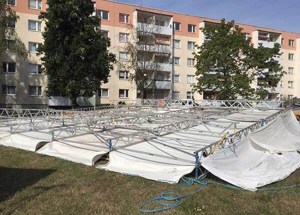 Metallgerüst und Zeltplane auf Rasen vor Mehrfamilienhaus bei Aufbauarbeiten.