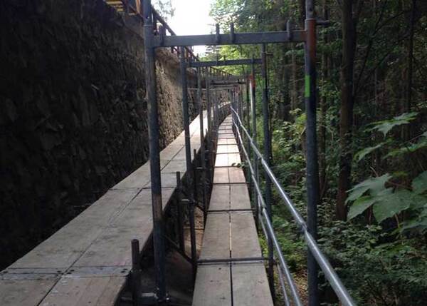 Gerüstbau mit Holzbohlen entlang einer Steinmauer im Wald für Bauarbeiten im Außenbereich.
