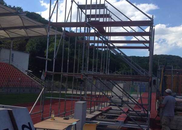 Gerüstkonstruktion im Stadion mit Zuschauertribüne und Sportplatz bei sonnigem Wetter.